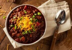 Bowl of chili topped with shredded cheddar cheese and chopped green onions, served with a spoon and napkin on a wooden table.